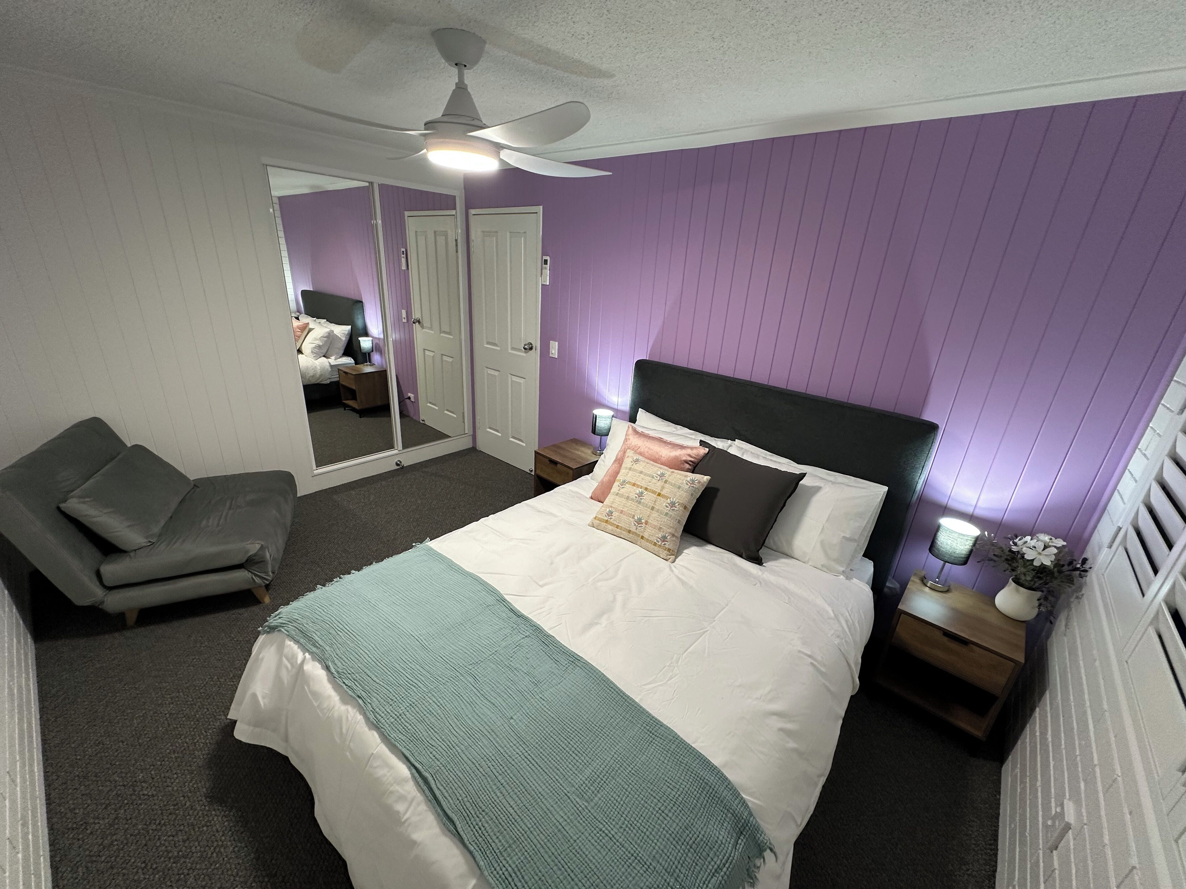 A bedroom with a bed, a chair, a ceiling fan, and a mirror.