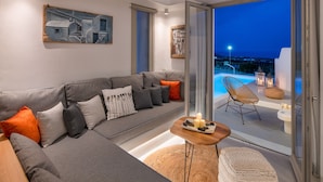 Ein modernes Wohnzimmer mit einem grauen Sofa, einem kleinen Holztisch mit Kerzen und Blick auf einen Pool und den Nachthimmel.