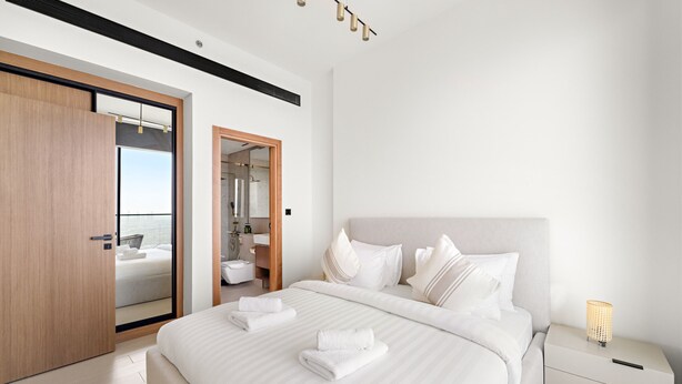 A modern hotel room with a large bed, a wooden wardrobe, a bathroom with a glass door, and a bedside table with a lamp. - Binghatti Gardenia - 3305 1BD (Dubai)