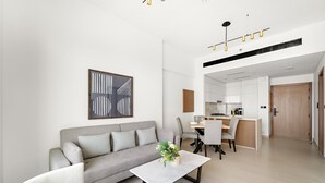 A modern living room with a grey sofa, a white coffee table, and a dining area with chairs and a kitchen in the background. - Binghatti Gardenia - 3305 1BD (Dubai)