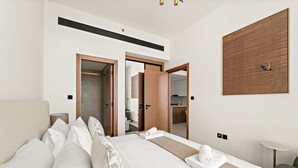 A modern hotel room with a large bed, a wooden headboard, a bedside table, a lamp, a mirror, and a view of the kitchen through an open door. - Binghatti Gardenia - 3304 1BD (Dubai)