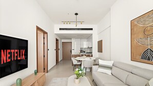 A modern living room with a flat-screen TV, a sofa, a coffee table, and a dining area with a kitchen in the background. - Binghatti Gardenia - 3304 1BD (Dubai)