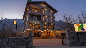Un hotel de varios pisos con fachada de piedra, balcones y un telón de fondo montañoso.