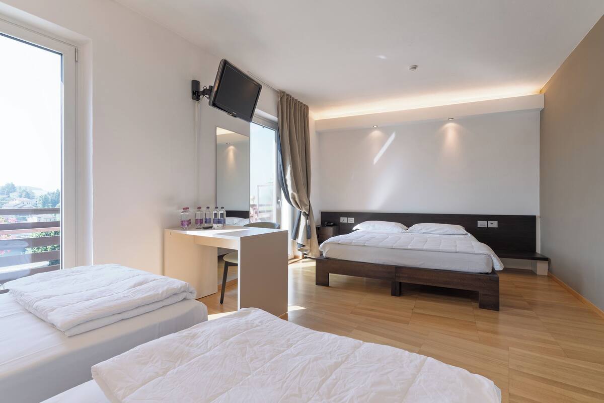 A modern hotel room with two beds, a desk, a TV mounted on the wall, and a large window offering a city view.