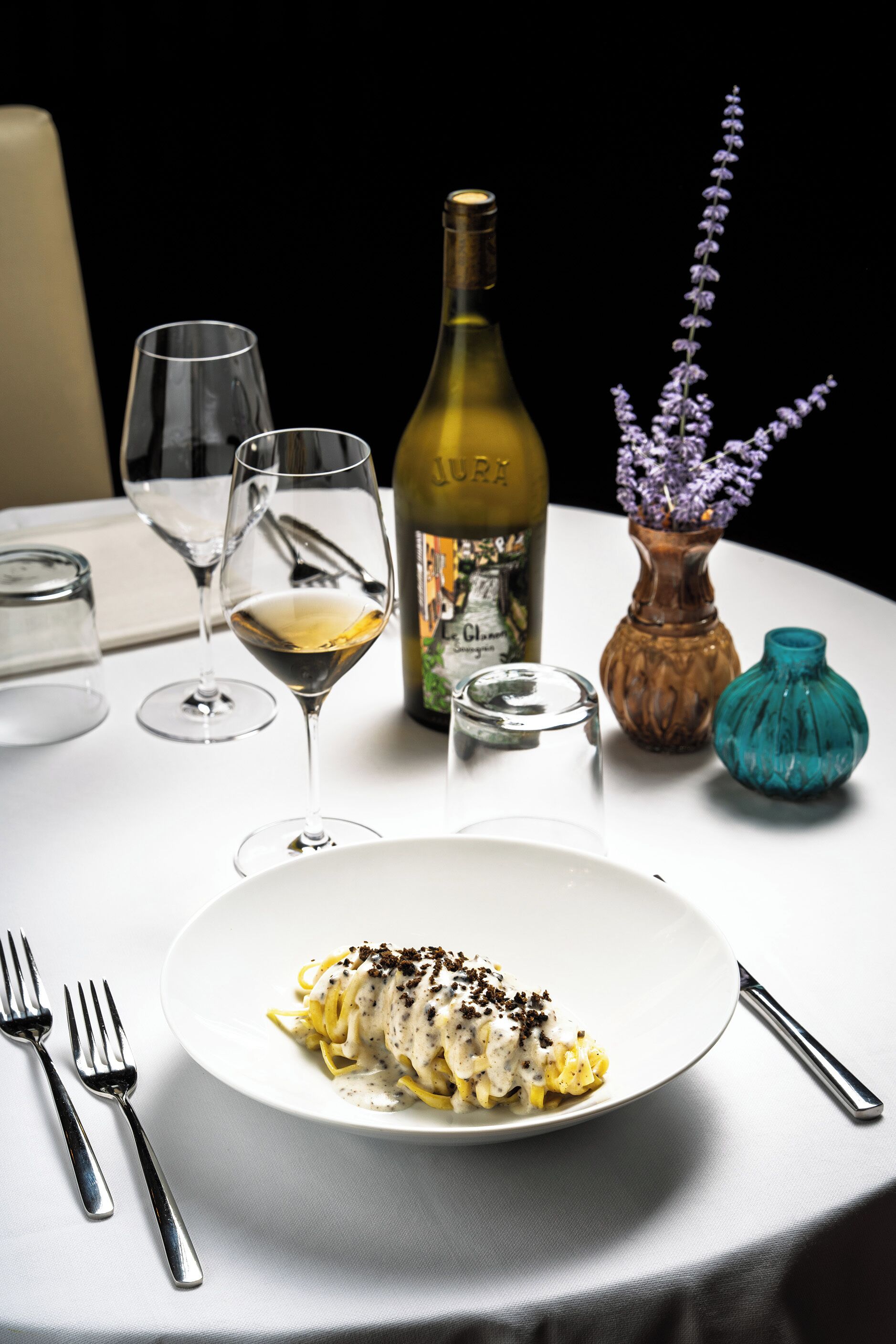 Una mesa de comedor con un plato de pasta, dos copas de vino, una botella de vino y dos jarrones con flores.