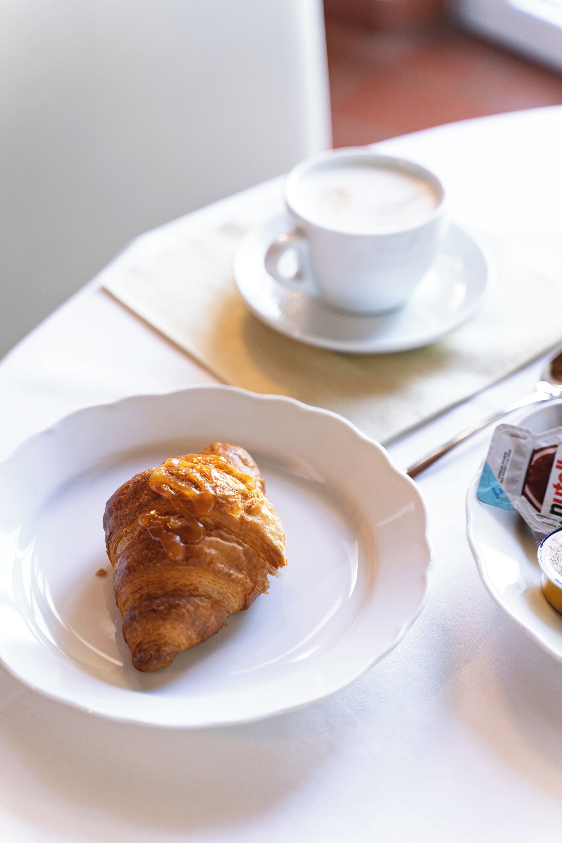 Un croissant sur une assiette blanche, avec une tasse de café sur une soucoupe en arrière-plan.