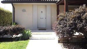 Ein Haus mit weißer Tür, einer kleinen Veranda und einem gepflegten Garten mit verschiedenen Sträuchern und Rasenfläche.