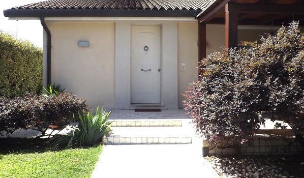 A house with a white door, a small porch, and a well-maintained garden with various shrubs and a lawn.