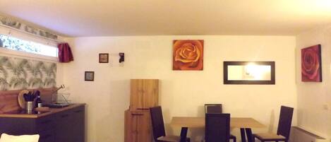 A compact dining area with a wooden table and four chairs, a small kitchen area with a sink and utensils, and a decorative wall with framed artwork.
