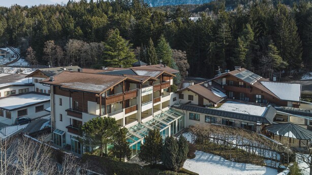 Ein Bergresort mit mehreren Gebäuden, darunter ein mehrstöckiges Haus mit Balkonen und einem Wintergarten, vor einer Kulisse aus schneebedeckten Gipfeln und dichtem Wald.