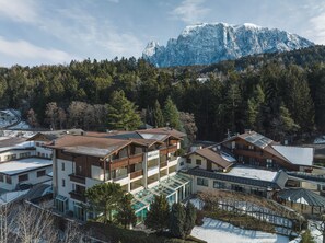 Une station de montagne comprenant plusieurs bâtiments, dont une structure à étages dotée de balcons et d’une véranda en verre, le tout situé dans un cadre spectaculaire, avec des sommets enneigés en toile de fond et une forêt dense.