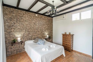 A bedroom with a stone wall, a bed with white linens, a wooden dresser, and a chandelier.