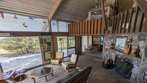 Amplia sala de estar con chimenea de piedra, asientos cómodos y ventanas grandes que ofrecen vistas al exterior.