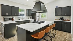 A modern kitchen with dark cabinetry, a large island with bar stools, a microwave, stove, and a large range hood. - Golden Dunes Stay Valpo | 5 BR, 3 King Beds | Game Area + Fire Pit + Deck Lounge (Valparaiso)