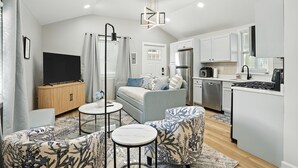 A modern apartment with a living room, kitchen, and dining area. It features a flat-screen TV, a sofa, a coffee table, and a dining table. The kitchen has a stove, oven, and dishwasher. - Golden Dunes Stay Valpo | 5 BR, 3 King Beds | Game Area + Fire Pit + Deck Lounge (Valparaiso)