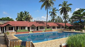 A pool area with a tiled deck, a red-roofed building, and palm trees.