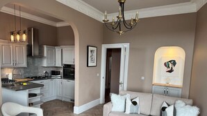 Een moderne woonkamer met een bank, een glazen salontafel en een eethoek, met een keuken op de achtergrond.