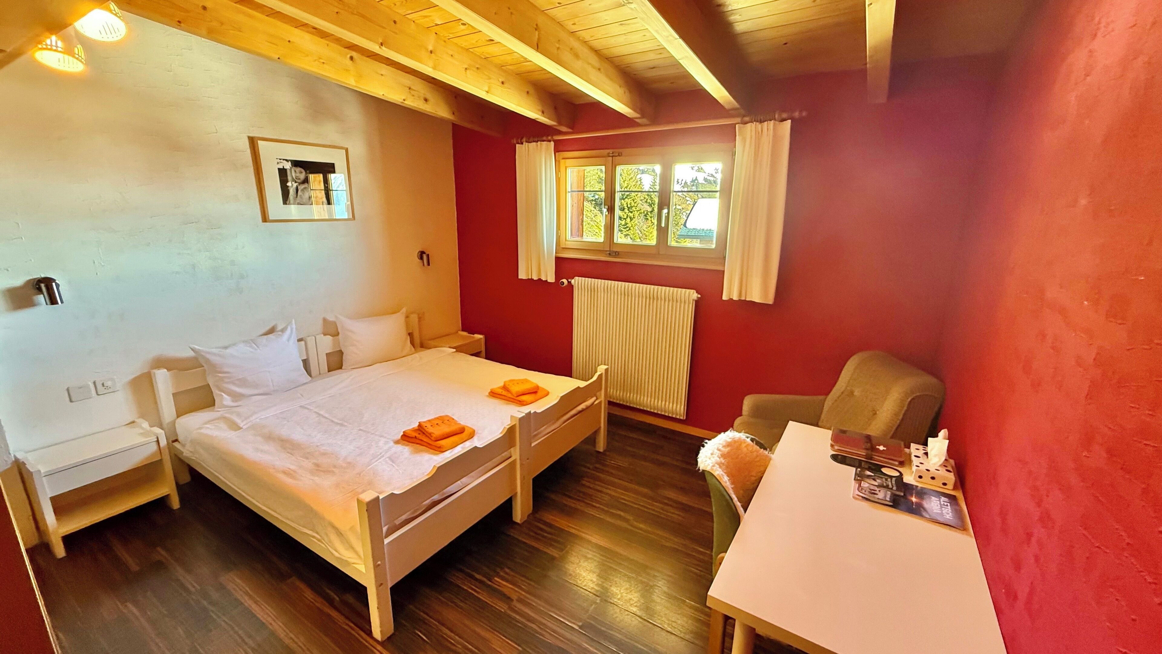 Quarto com uma cama, uma cadeira, uma mesa, uma janela com cortinas, um radiador e um quadro na parede.
