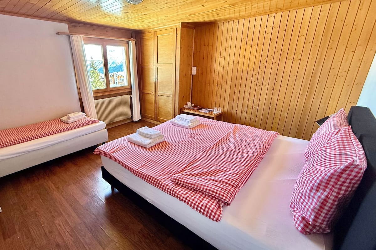 Una habitación con dos camas, una más grande y otra más pequeña, ambas con ropa de cama a cuadros rojos y blancos. Hay una mesita con una lámpara y una ventana con cortinas.