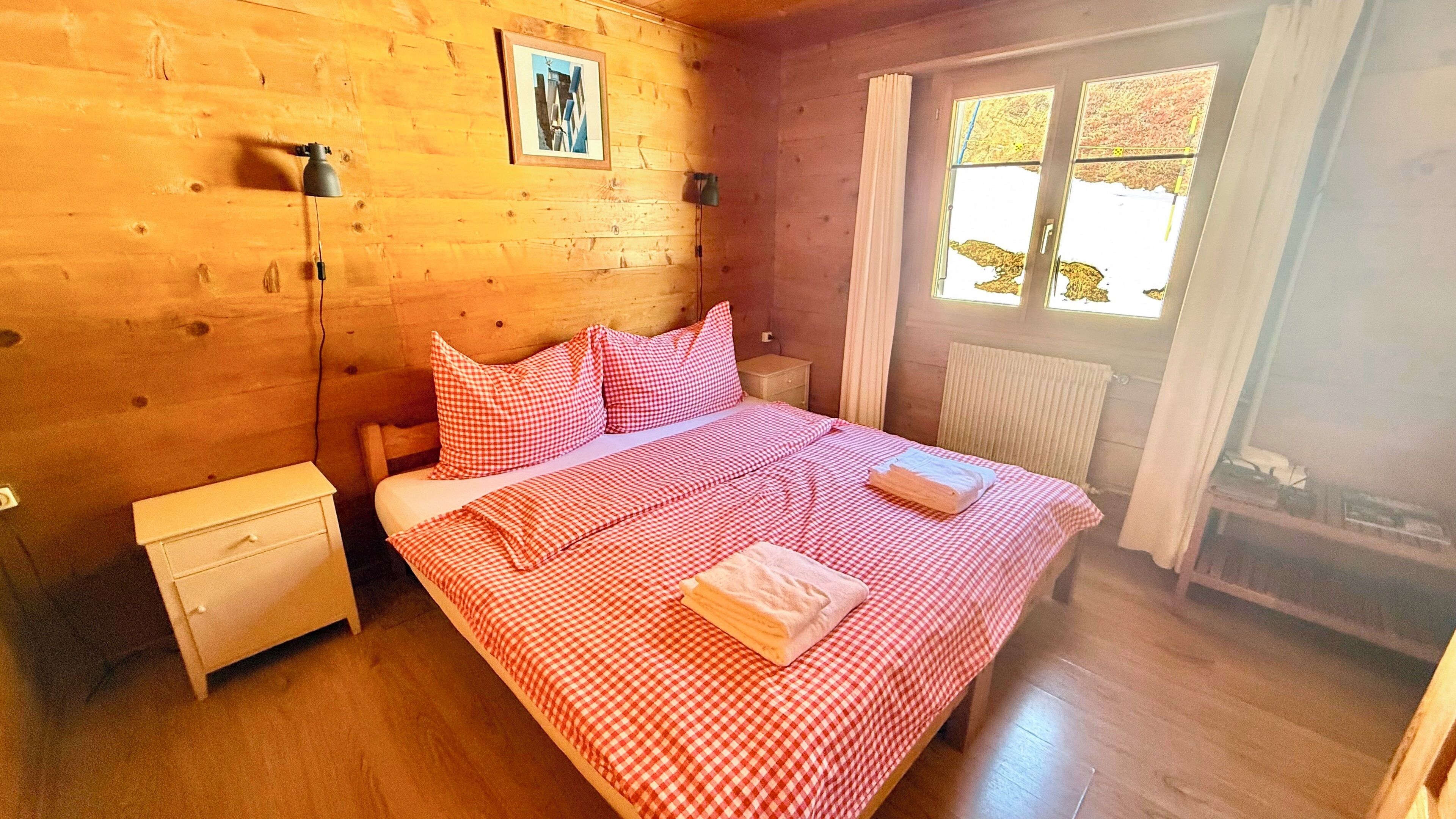A wooden room with a bed, a nightstand, a lamp, and a window with curtains.