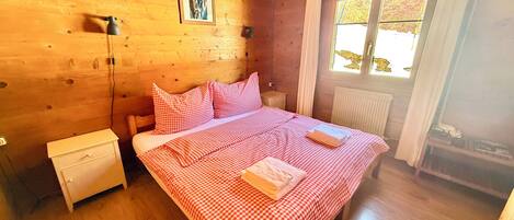 A wooden room with a bed, a nightstand, a lamp, and a window with curtains.