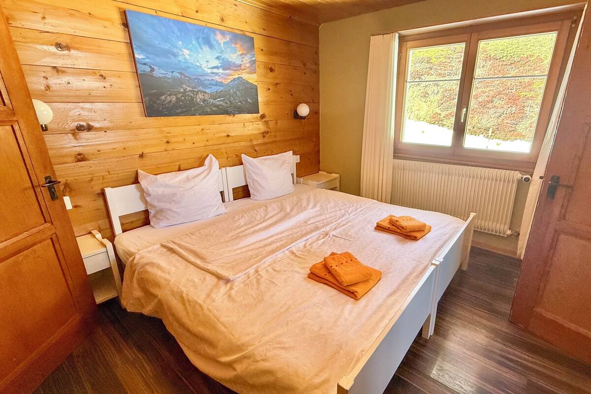 Un dormitorio con pared de madera, una cama con ropa blanca, un cuadro sobre la cama y una ventana con vistas a árboles.