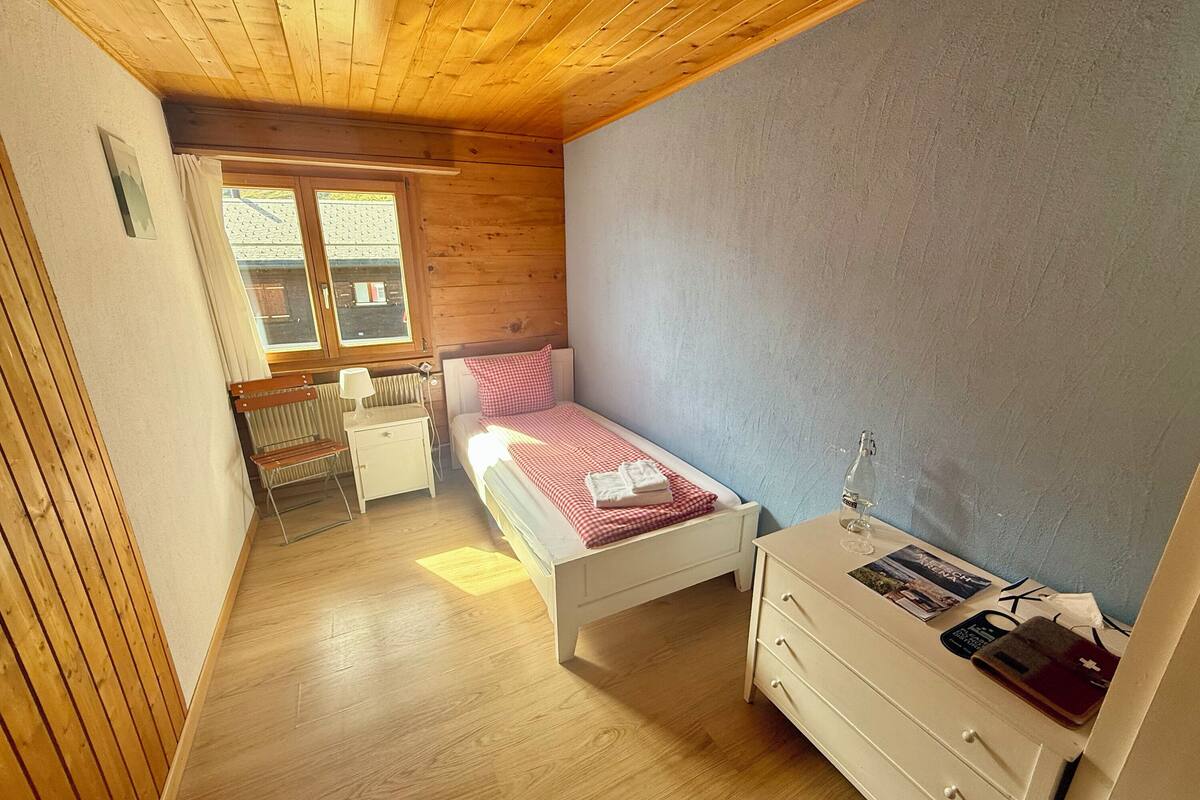 Un dormitorio con techo de madera, una cama con colcha a cuadros rojos, una mesita de noche con lámpara y una ventana con cortinas.
