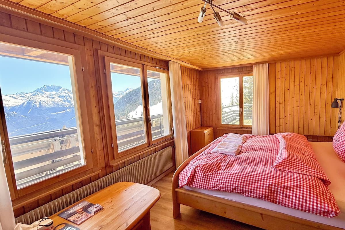 Habitación de cabaña de madera con cama, una mesa y ventanas amplias que dan a vistas de la montaña.