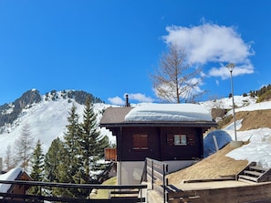 A wooden cabin with a snow-covered roof, surrounded by snow-covered mountains and trees.
