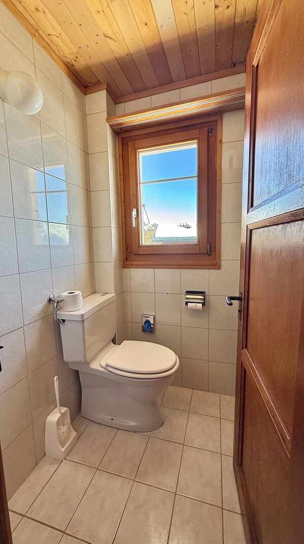 A small bathroom with a toilet, a window, and a wooden door. - Berghaus Toni Hotel und Restaurant (Riederalp)