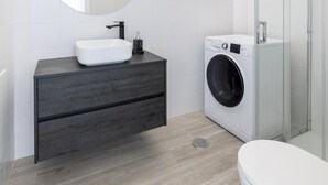 A modern bathroom with a white sink, dark wood vanity, a round mirror, a washing machine, and a glass shower enclosure.