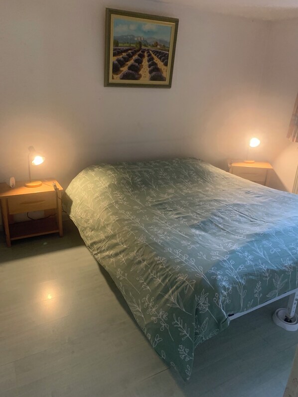Un dormitorio con colcha estampada de flores verdes, dos lámparas de noche, una mesita de noche y un cuadro con paisaje en la pared.