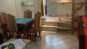 A dining area with a table and chairs, a kitchen in the background, and a staircase leading to an upper level.