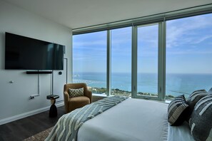 A bedroom with a large bed, a TV mounted on the wall, and a view of the ocean through large windows.