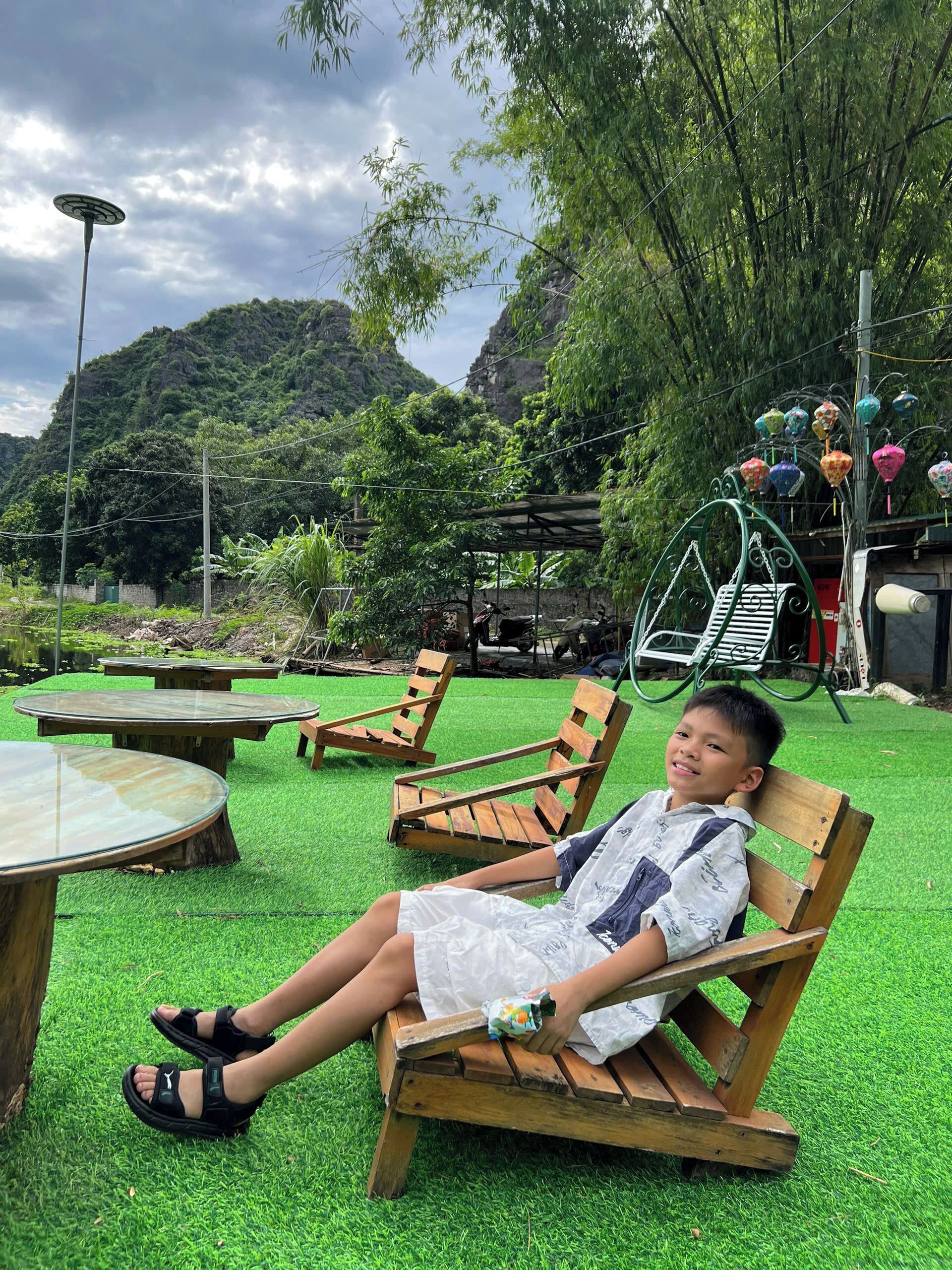 A child sitting on a wooden chair in a garden with artificial grass, surrounded by tables and chairs.
