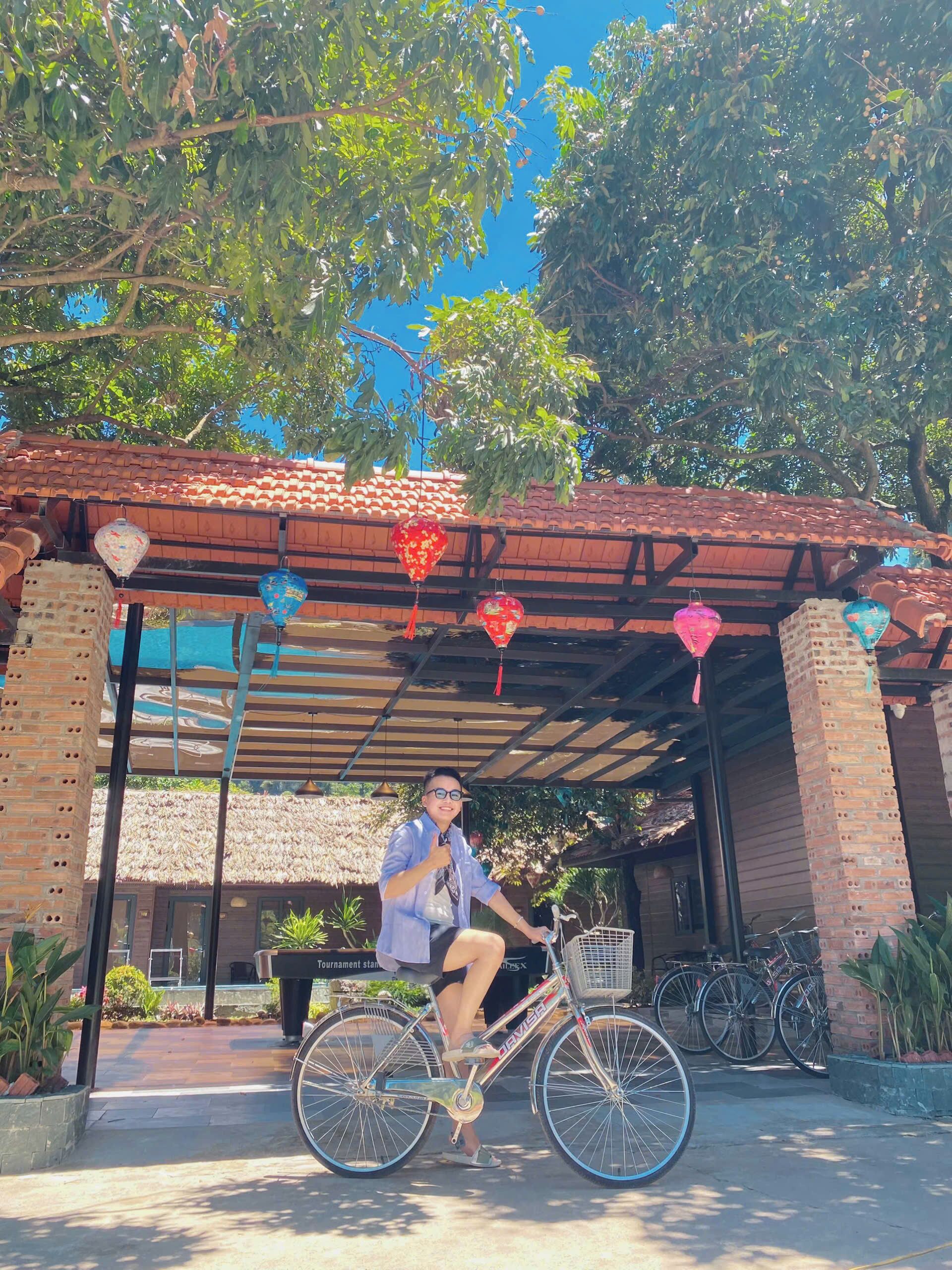 Una persona sentada en una bicicleta bajo un espacio cubierto, con un muro de ladrillo y un techo de paja al fondo.