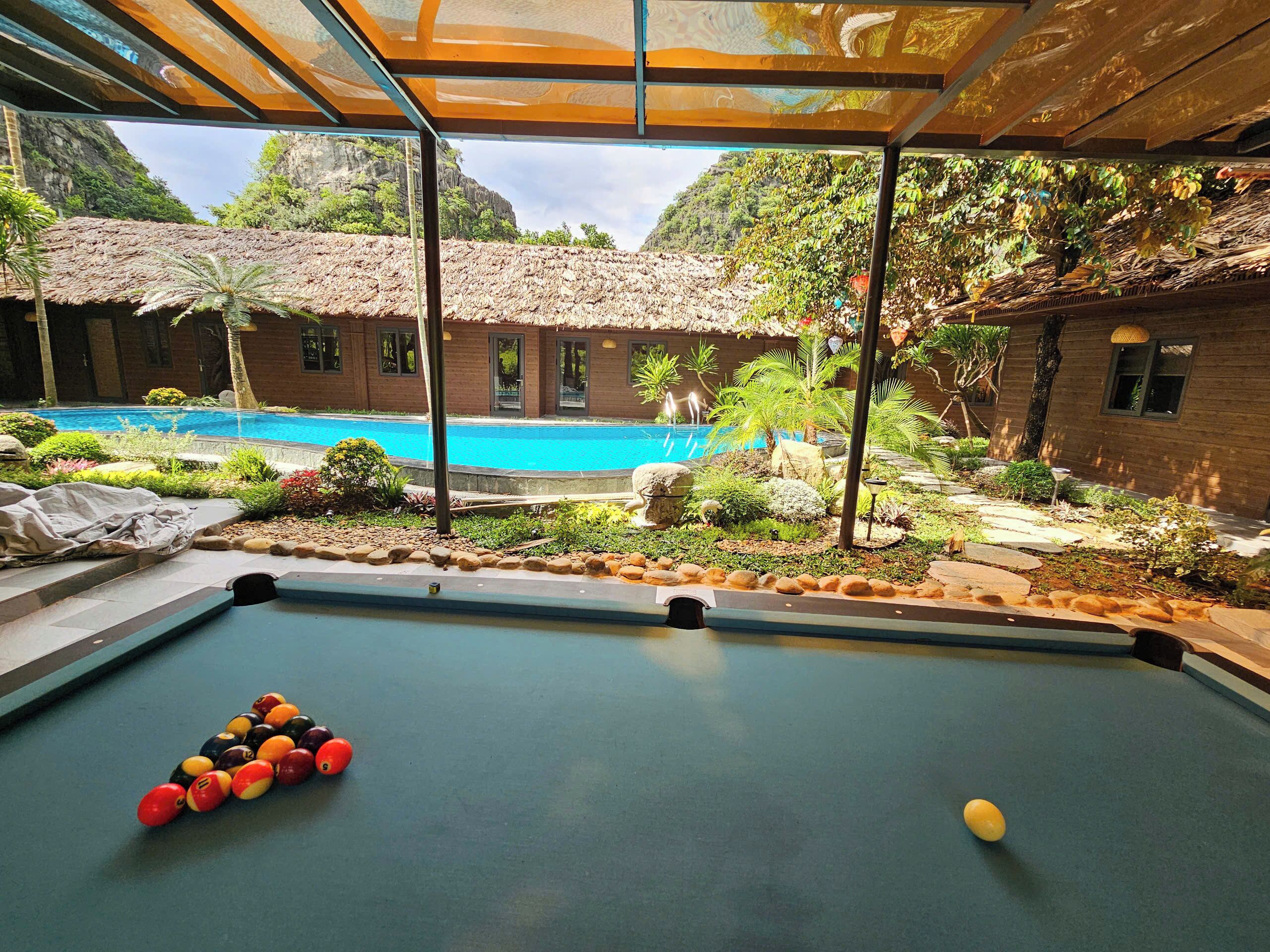 A pool table with billiard balls, a pool, and a building with a thatched roof in the background.