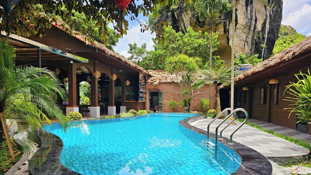 A swimming pool with a surrounding deck, a bar area, and a natural backdrop of trees and rock formations. - An Riverside Ecolodge (Tay Hoa Lu)
