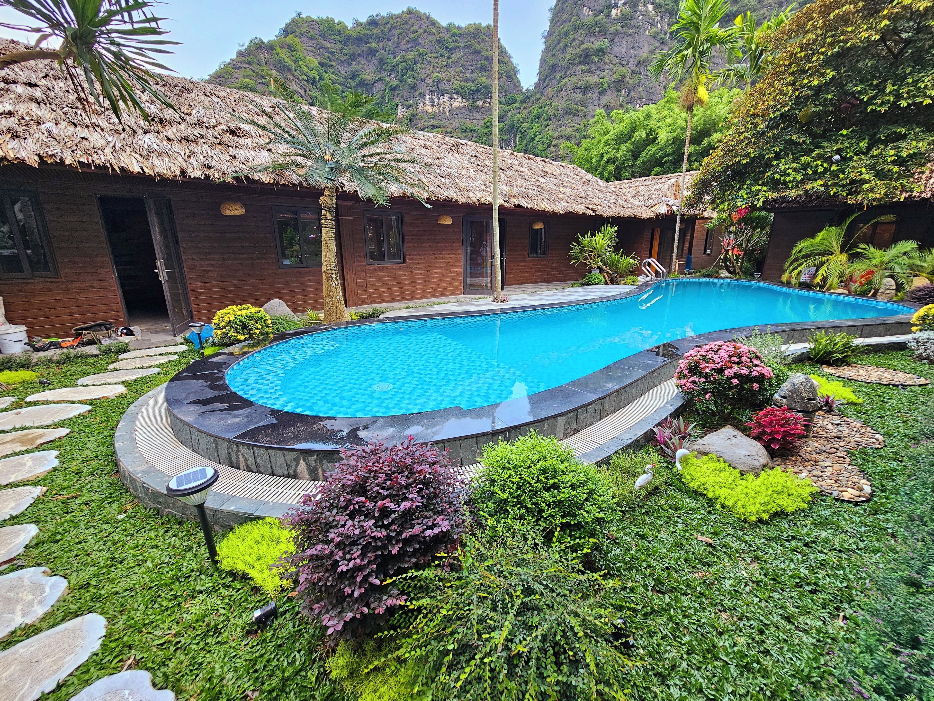 Une piscine entourée d’une végétation luxuriante, un bâtiment en bois avec un toit de chaume et, en arrière-plan, un paysage montagneux.
