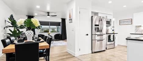 A modern kitchen with stainless steel appliances, a dining area with a wooden table and black chairs, and a view through a glass door to an outdoor area.