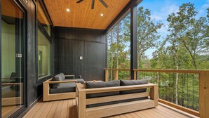 A modern outdoor seating area with wooden benches and cushions, large windows, and a view of trees. - New Lake House on Lake Adger near TIEC, Vineyards, Orchards, Biltmore and more (Mill Spring)