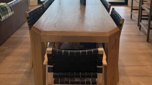 A large wooden dining table with black cushioned chairs, a sofa, and a view of a forest through large windows. - New Lake House on Lake Adger near TIEC, Vineyards, Orchards, Biltmore and more (Mill Spring)