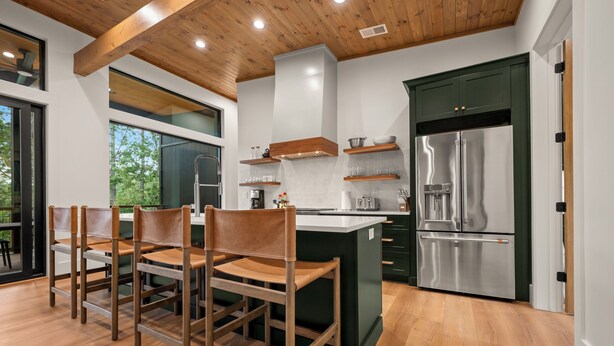 A modern kitchen with wooden chairs, a stainless steel refrigerator, and a large window overlooking greenery. - New Lake House on Lake Adger near TIEC, Vineyards, Orchards, Biltmore and more (Mill Spring)