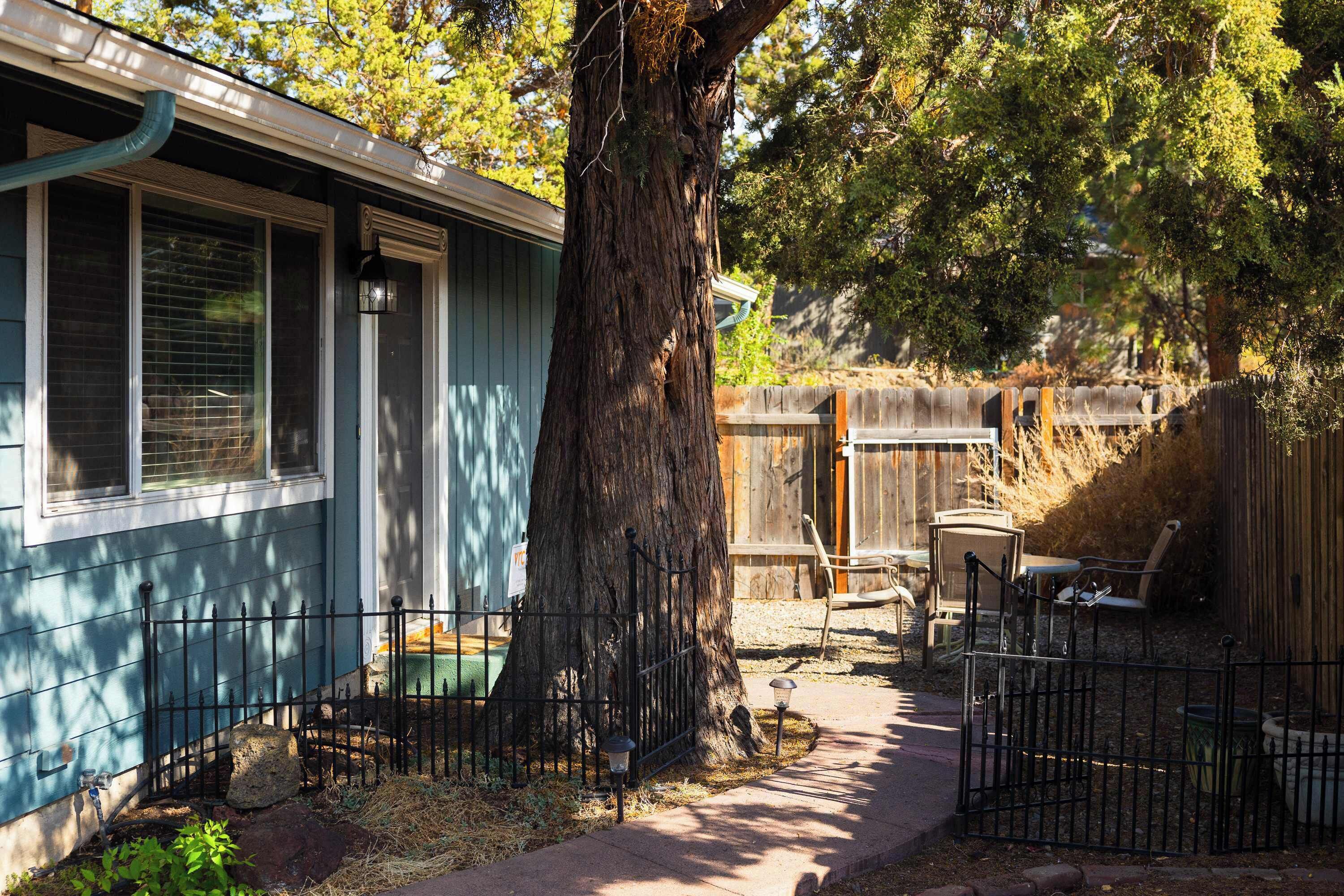 A house with a large tree in front, a fenced backyard, and outdoor furniture.