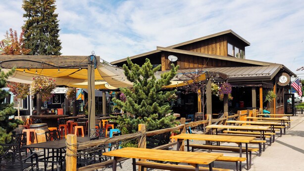 An outdoor seating area with wooden tables and chairs, a covered patio, and a building with a clock and American flag. - 10min to Downtown Bend Chic 2bdr (Bend)