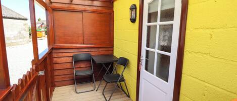 Un área de patio techada con una mesa y sillas de madera, una puerta con paneles de vidrio esmerilado y una pared amarilla.