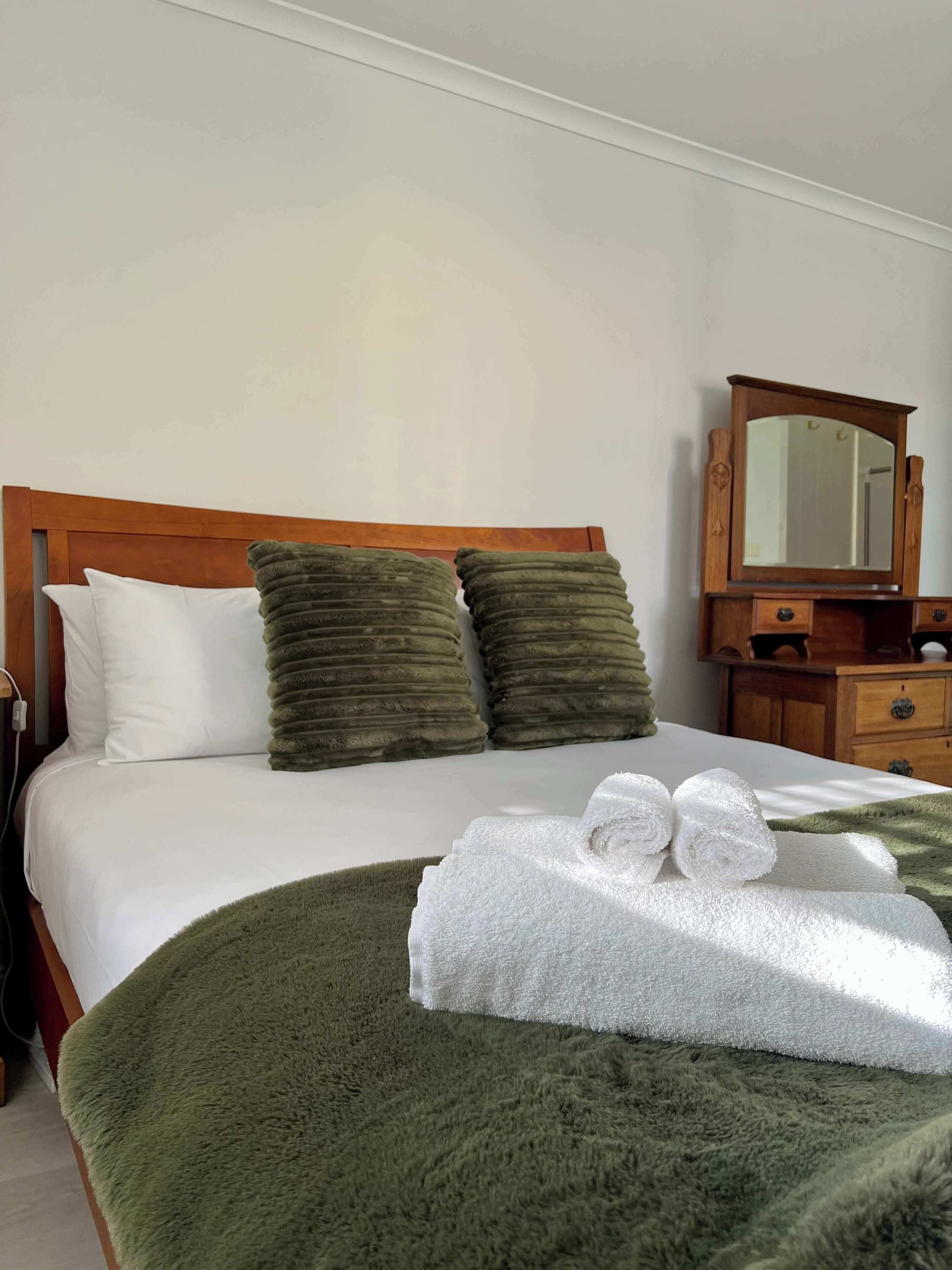 Une chambre d’hôtel avec un lit, des oreillers, un plaid vert, une table de chevet en bois, un miroir et une serviette.