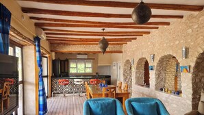 A cozy living room with a fireplace, a dining area with a table and chairs, and a kitchen in the background. - Riad Ahlam villa with swimming pool surrounded by argan trees (Essaouira)