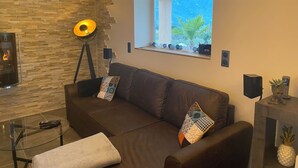 A modern living room with a stone wall, a sofa with patterned cushions, a floor lamp, and a window with a view of greenery. - Villa Ghjuvan (Peri)