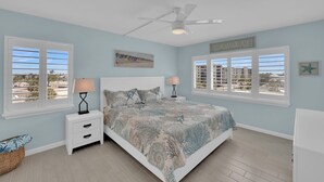 Ein Schlafzimmer mit einem Bett, zwei Nachttischen mit Lampen, einem Deckenventilator und großen Fenstern mit Blick auf den Strand.